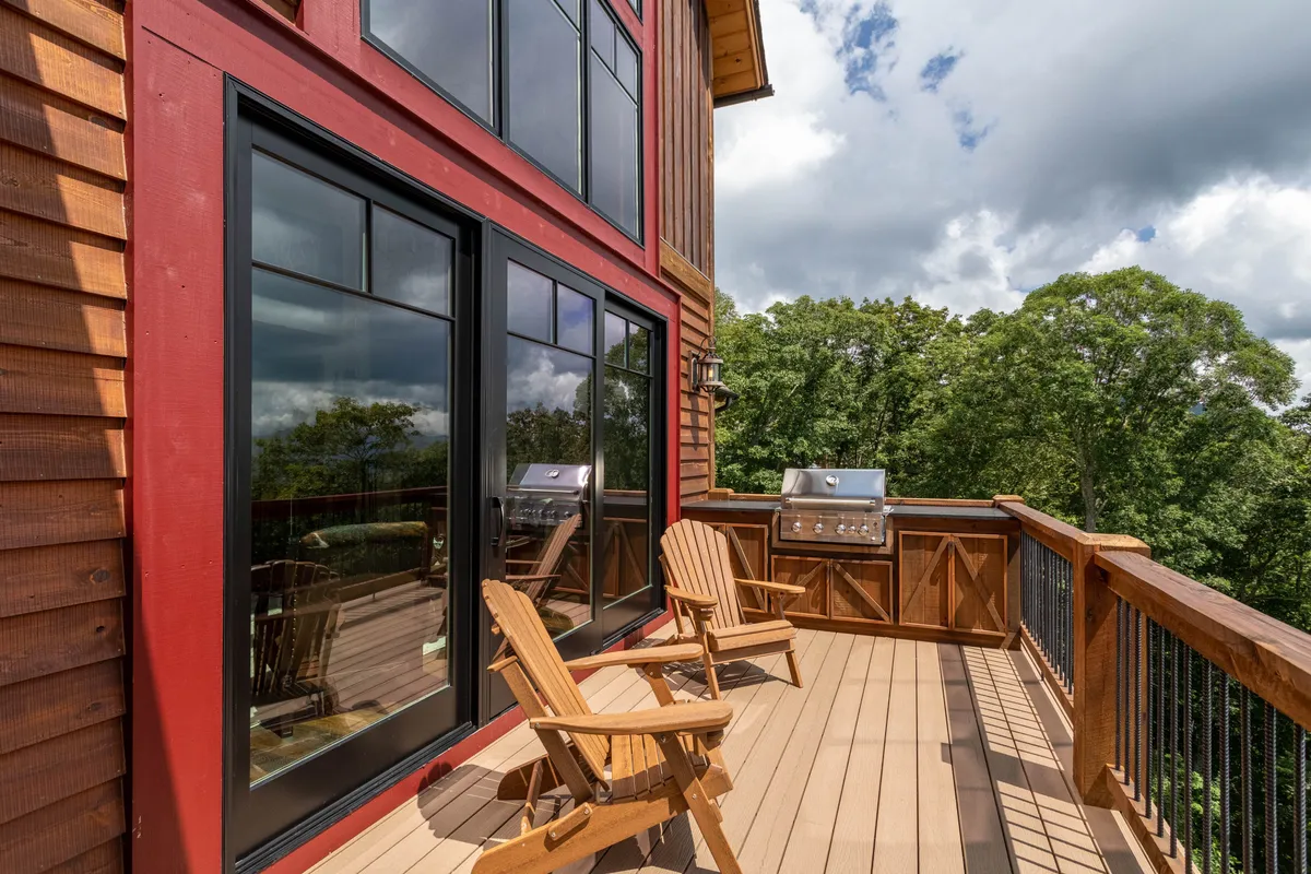 An inviting wooden deck featuring Adirondack chairs and a grill, with large windows reflecting the surrounding landscape. The relaxed setup suggests opportunities for both social gatherings and solitary moments, from barbecuing to simply enjoying the sights and sounds of nature.