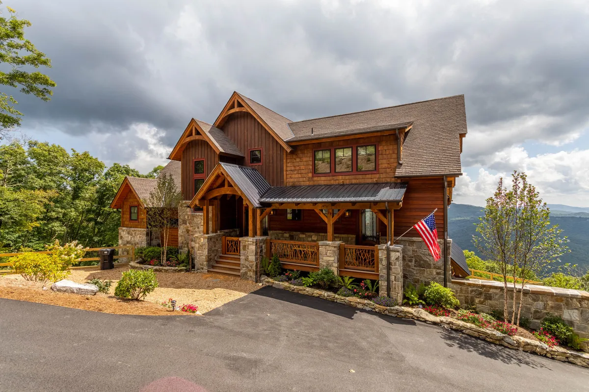 This image shows the inviting exterior of the property, featuring a warm wooden entrance, stone facade, and flag pole. The porch looks perfect for relaxing while enjoying views of the rolling hills and surrounding landscape. The setting sun hints at beautiful evening ambiance.