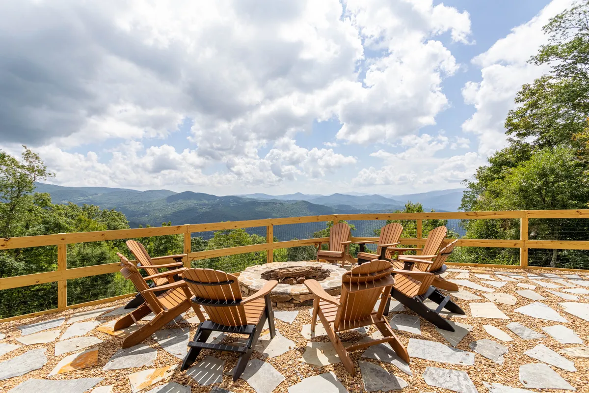 A cozy fire pit area surrounded by classic Adirondack chairs, set against a stunning mountain view. The space is perfect for gathering, sharing stories, or stargazing under the open sky. A wooden railing frames the deck, emphasizing safety and intimacy.