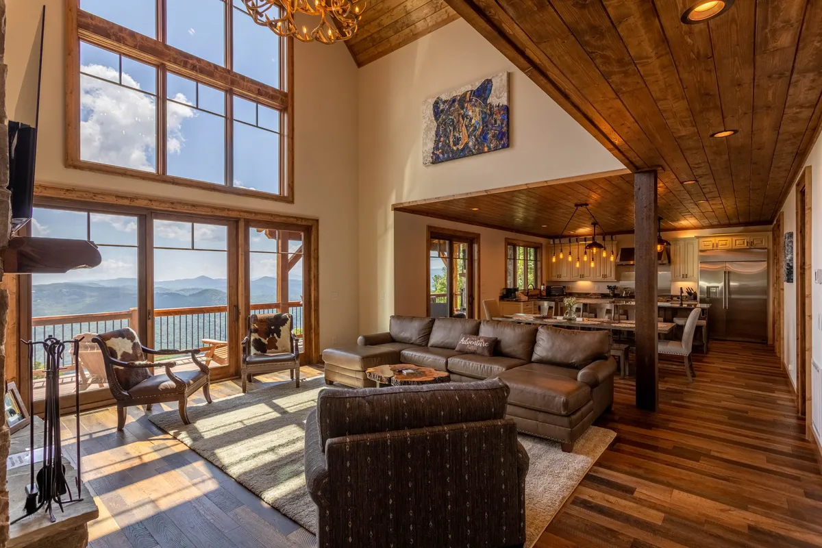 This image showcases a spacious great room with large windows offering sweeping mountain views. A comfortable sectional sofa is arranged to enjoy both the view and the stone fireplace. The room is highlighted by warm lighting and tasteful artwork, creating an inviting space for gatherings and relaxation.