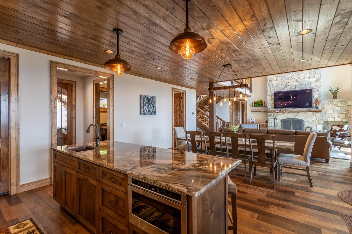 The image features a gourmet kitchen with a large kitchen island, adjacent dining table, stone fireplace, and wooden ceiling. The space is well-lit and designed for both cooking and socializing, embodying a communal atmosphere ideal for family feasts and group gatherings.