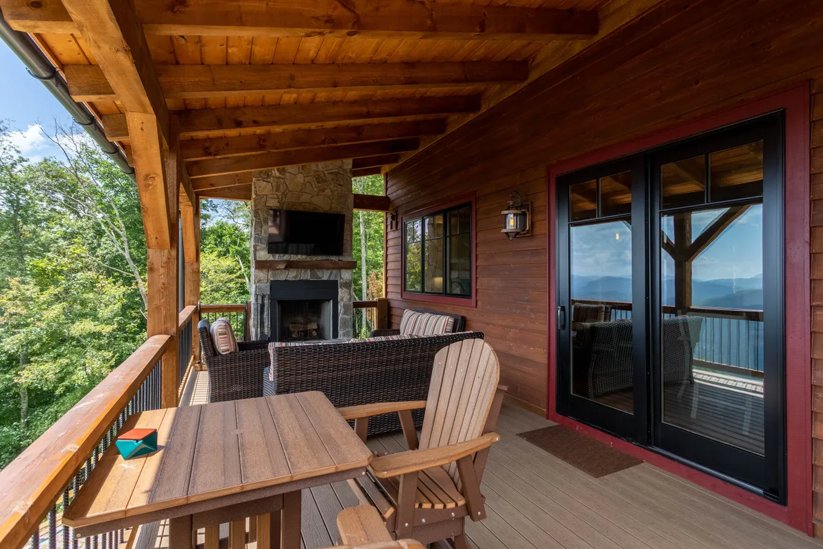 An inviting covered deck is shown with a stone fireplace, comfortable outdoor seating, and a backdrop of trees and mountains. The mood is serene and cozy, suggesting a perfect spot for al fresco dining or sunset cocktails year-round.