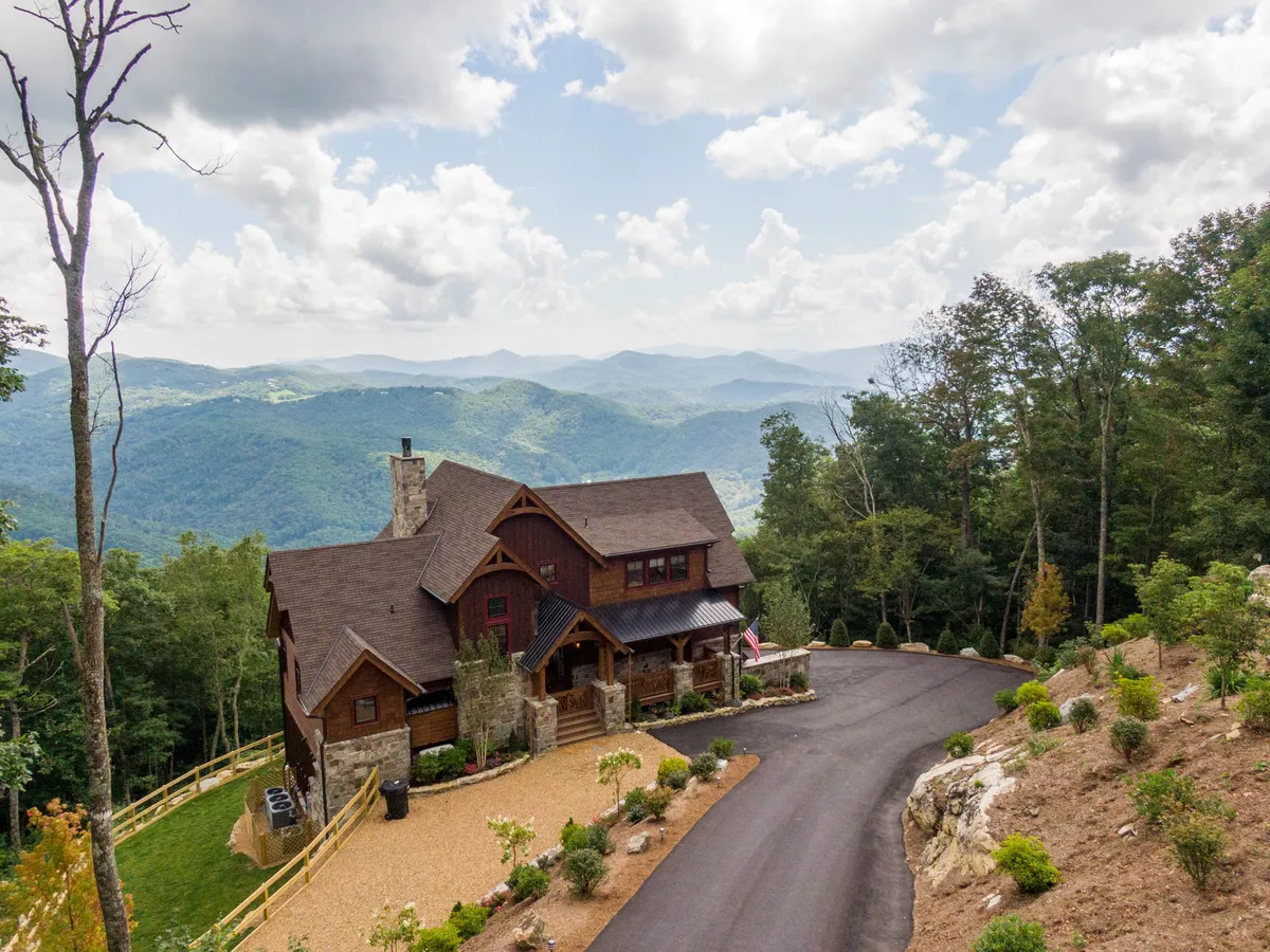 The image displays a striking, newly built mountain home set against the breathtaking backdrop of rolling Blue Ridge vistas. The house sits at the top of a gentle driveway, surrounded by lush gardens and native landscaping, with a spacious porch perfectly positioned for sunset viewing. The setting radiates peace and invites outdoor gatherings with family and friends, while the expansive mountain scenery promises awe-inspiring mornings and cozy evenings under the stars.