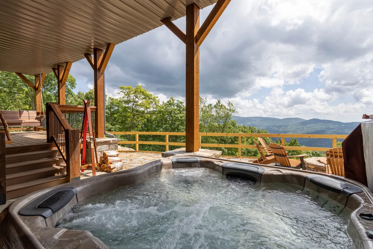 A serene exterior shot featuring a luxurious hot tub set on a wooden deck, overlooking sweeping mountain views. Rock formations and deck chairs add to the tranquil ambiance, while the hot tub is positioned for optimal enjoyment of the surrounding landscape.